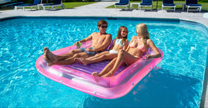 Three people relaxing on a pink inflatable raft in a pool.